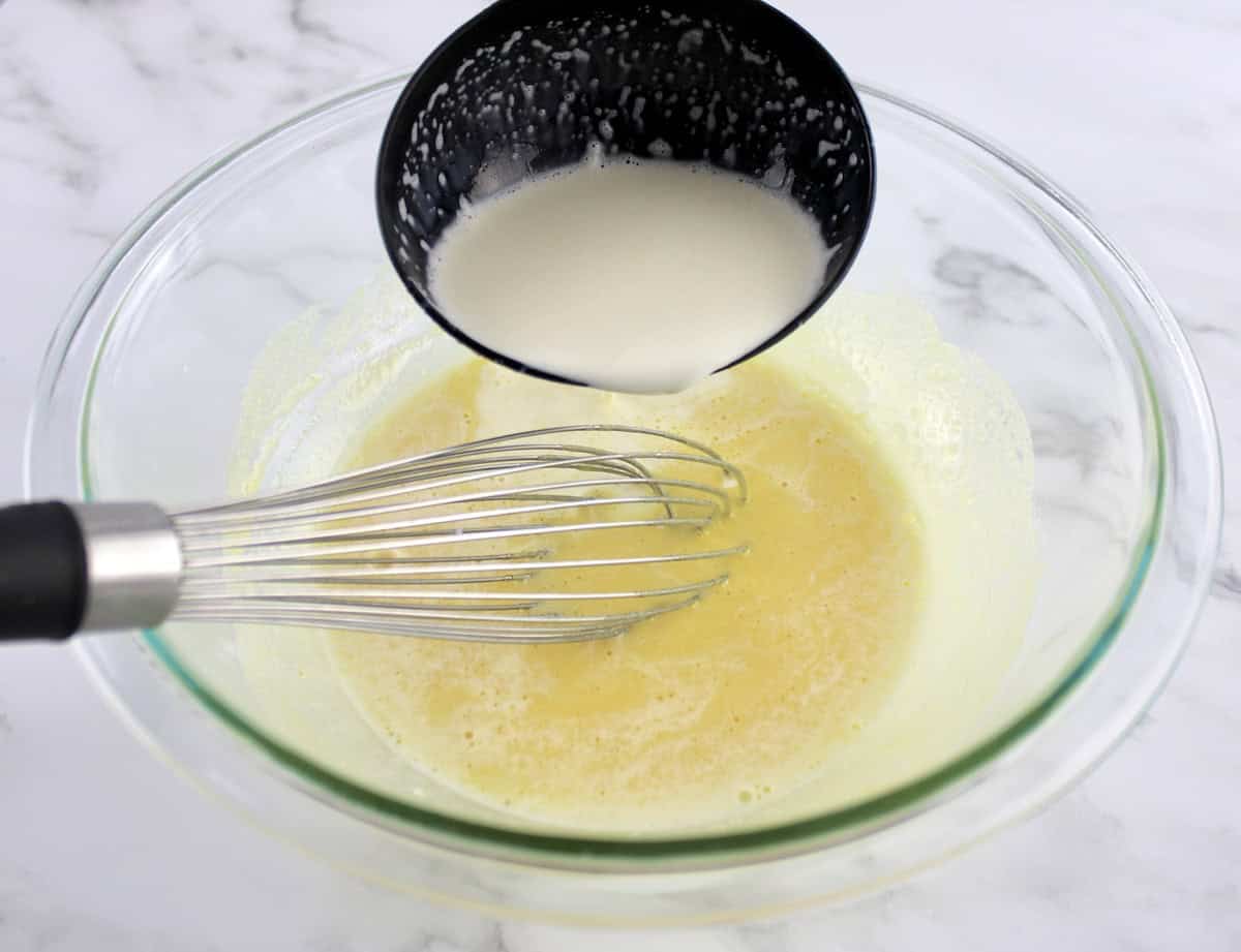 cream being ladled into glass bowl with egg yolks and whisk