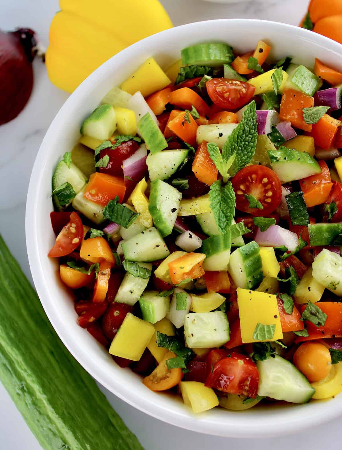 Israeli Salad in white bowl with mint sprig in center and veggies in background