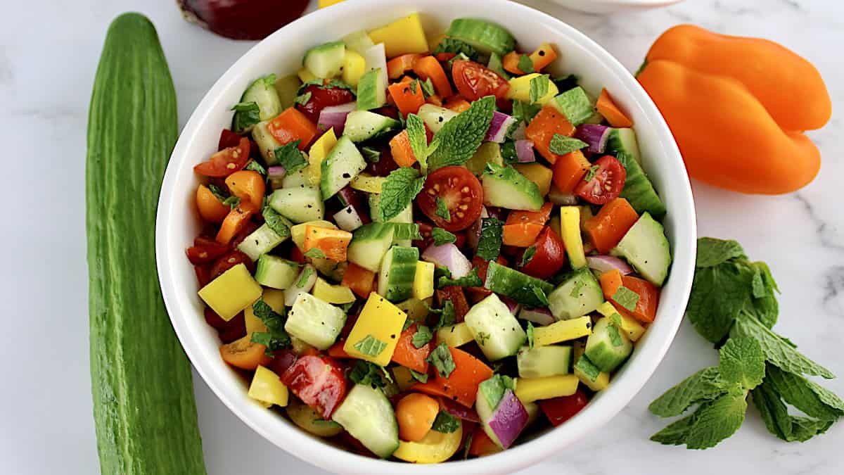 overhead view of Israeli Salad in white bowl with veggies and mint in background