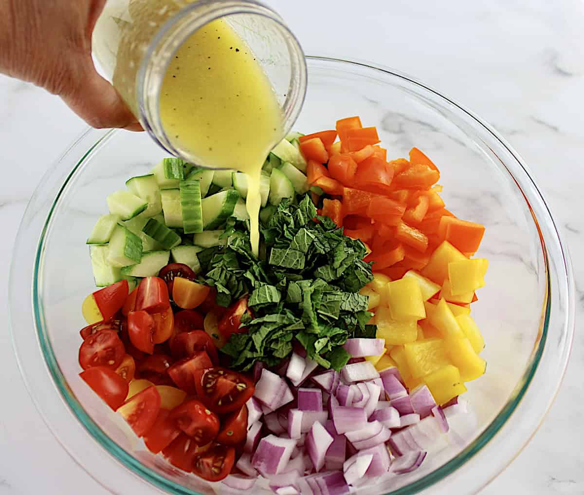 Israeli Salad ingredients in groups in glass bowl unmixed with dressing being poured over top