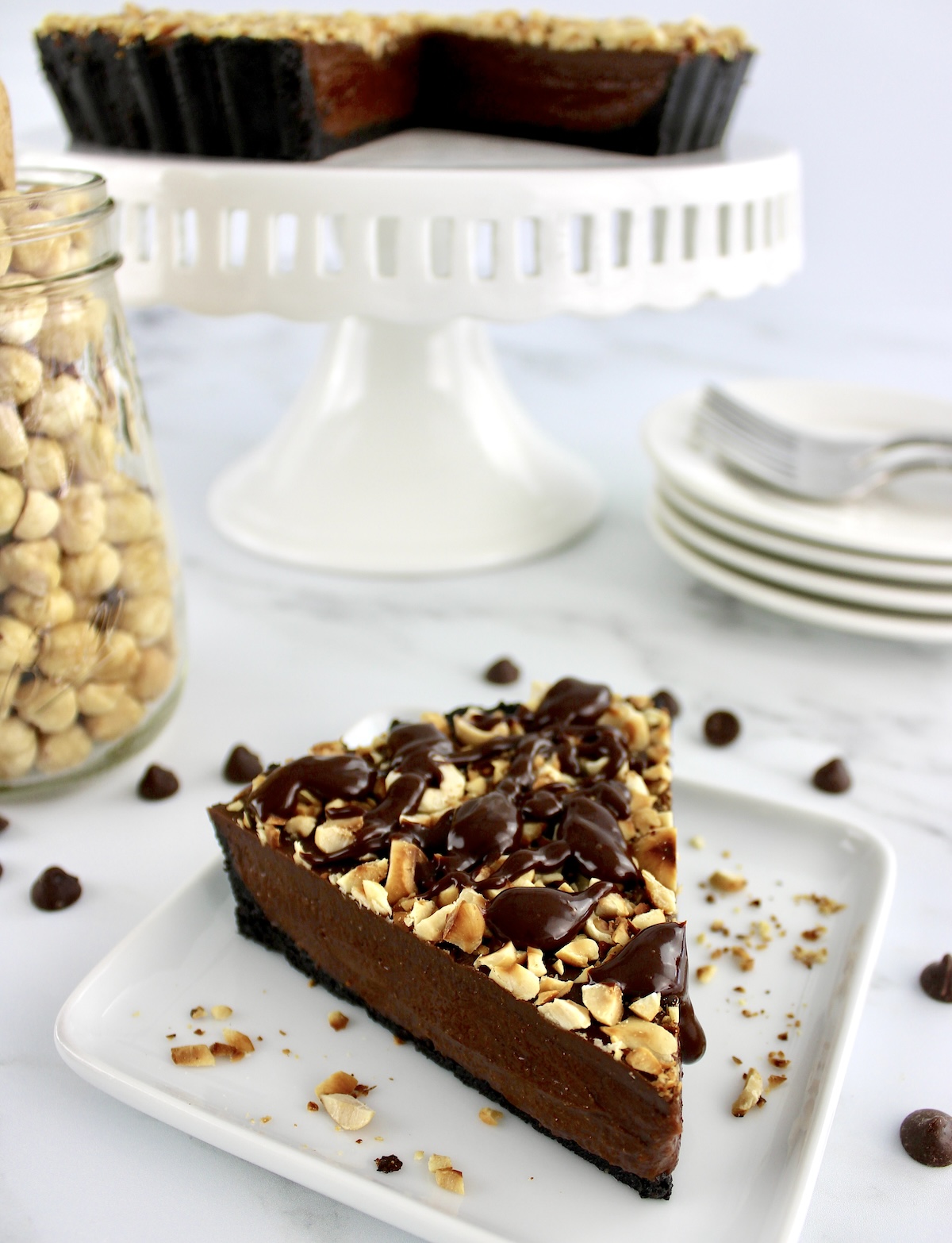 Chocolate Hazelnut Tart slice on white plate with white cake stand and hazelnuts in back