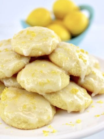 Lemon Ricotta Cookies stacked up on white plate with lemons in background