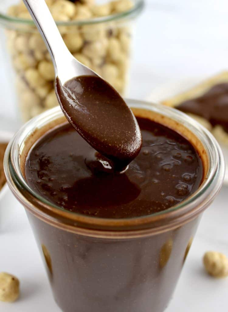 Sugar-Free Nutella being spooned out of glass jar with hazelnuts in glass jar in back