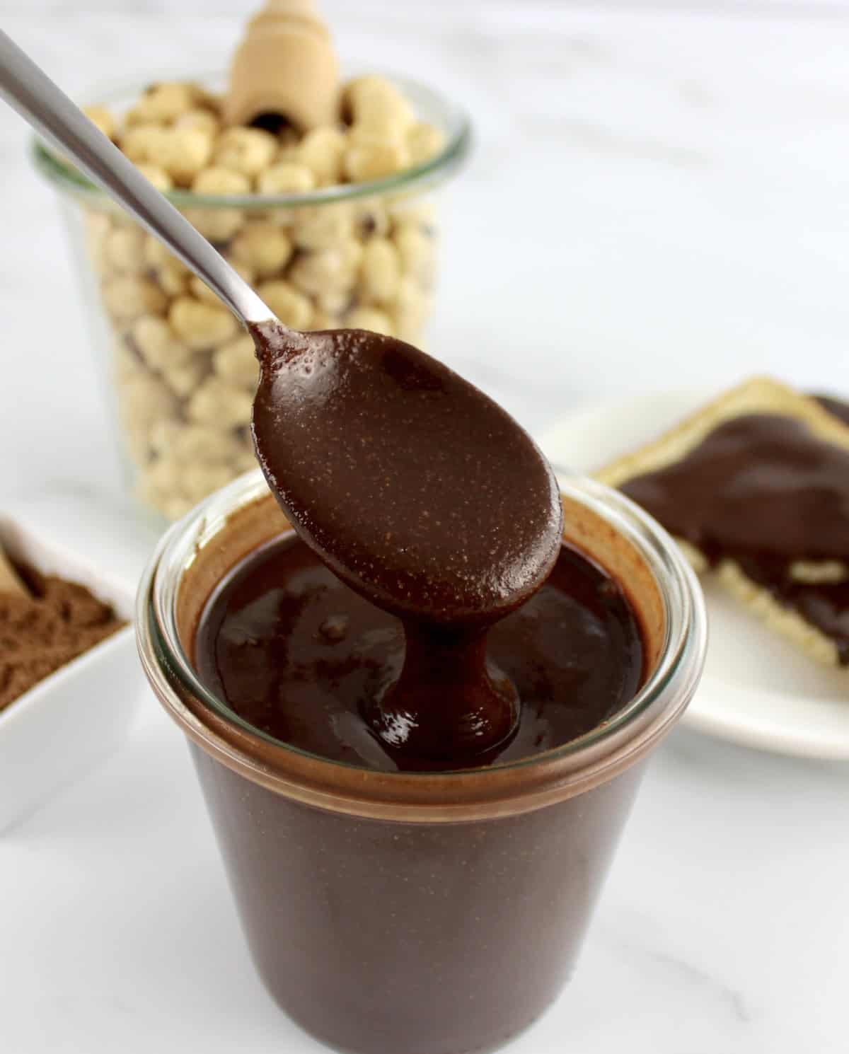 Sugar-Free Nutella being spooned out of glass jar with hazelnuts in glass jar in back