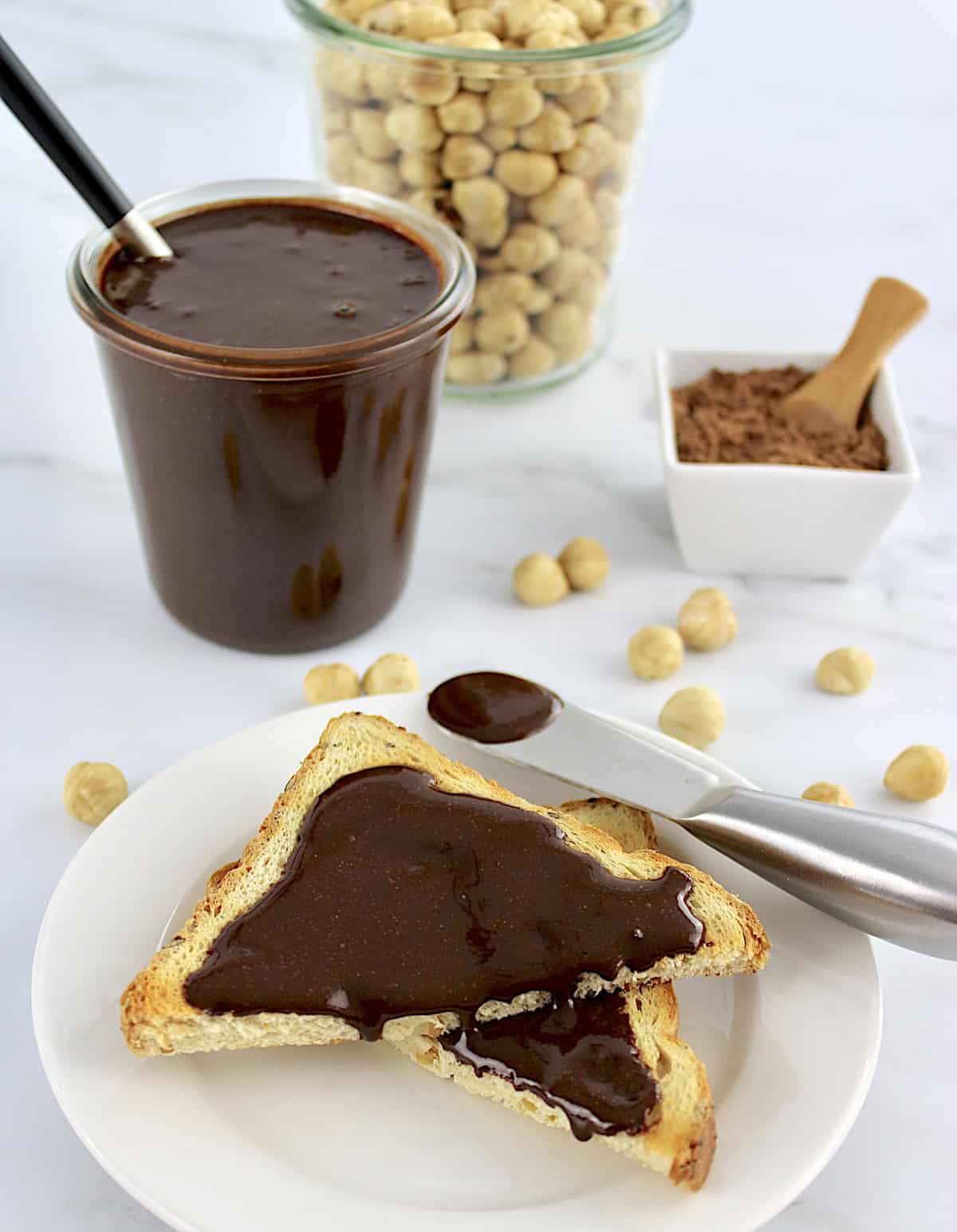 Sugar-Free Nutella on 2 cut pieces of toast on white plate with nutella in glass jar in back