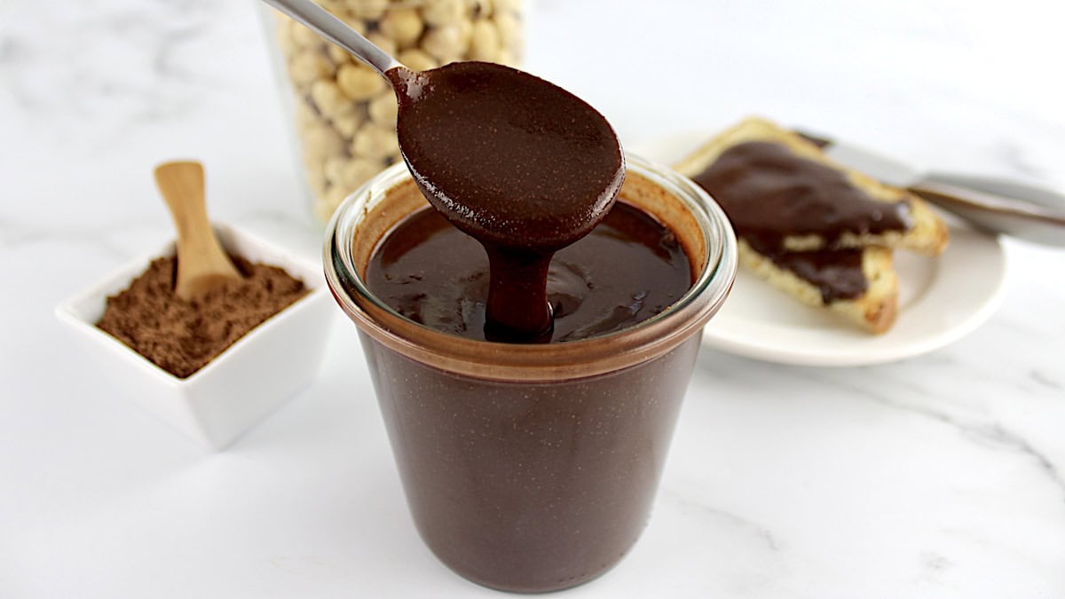 Sugar-Free Nutella being spooned out of glass jar with hazelnuts in glass jar in back