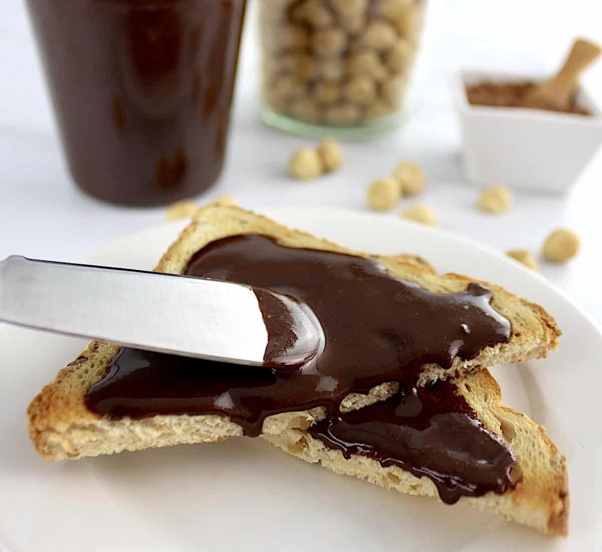 closeup of spreading nutella on toast with nutella and hazelnuts in back