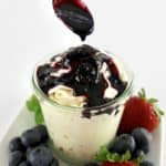 Keto Blueberry Sauce being spooned over cheesecake fluff in glass jar with berries around