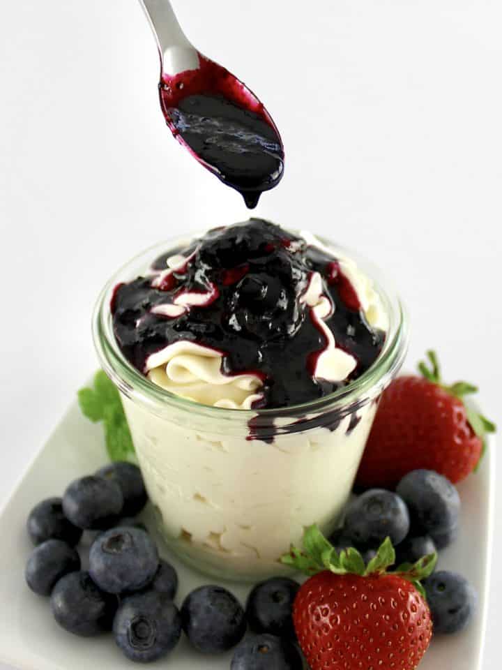 Keto Blueberry Sauce being spooned over cheesecake fluff in glass jar with berries around