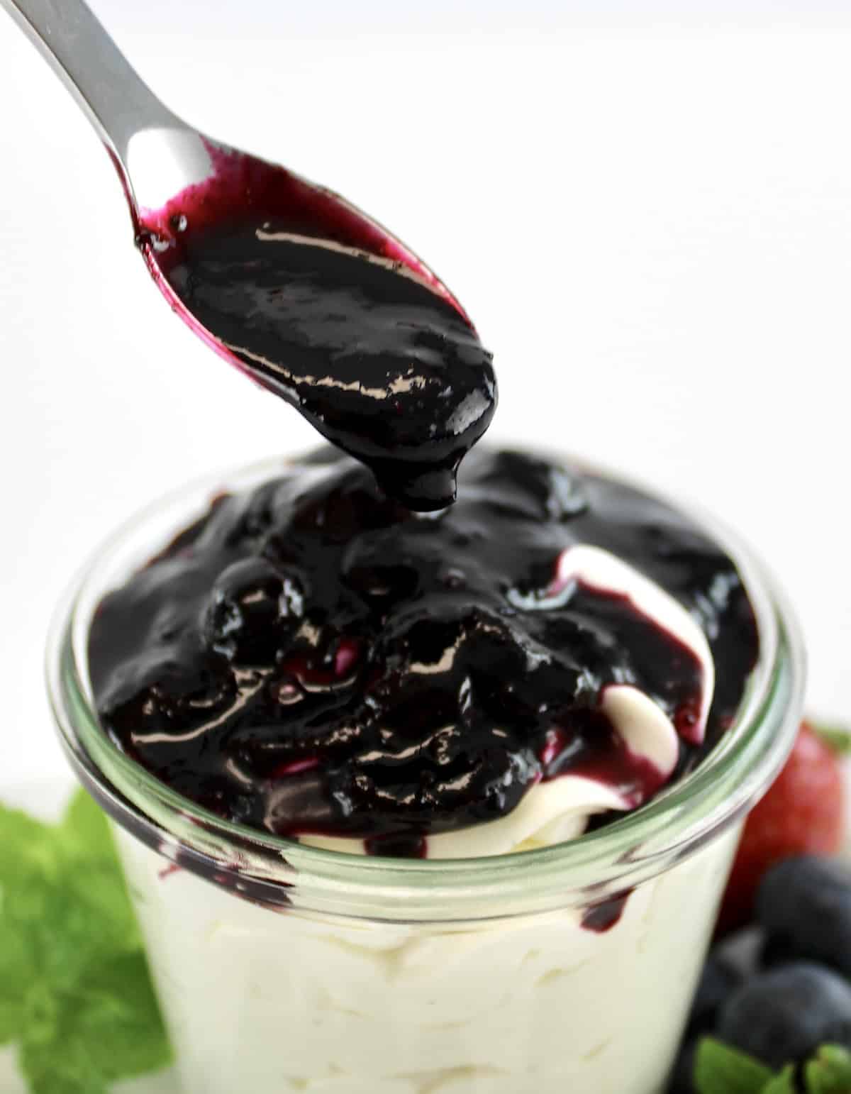 Keto Blueberry Sauce being spooned over cheesecake fluff in glass jar with berries around