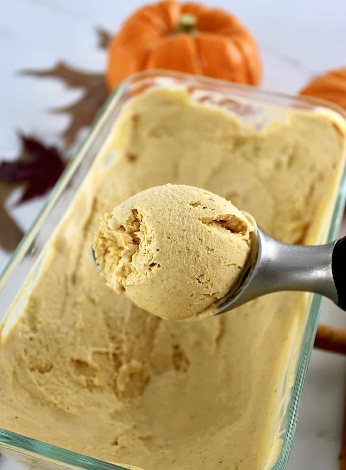 No-Churn Pumpkin Ice Cream being scooped out of glass container with pumpkin in back