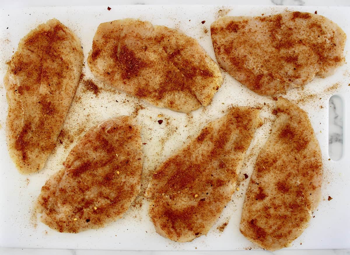 6 pieces of raw chicken breast with taco seasoning on white cutting board