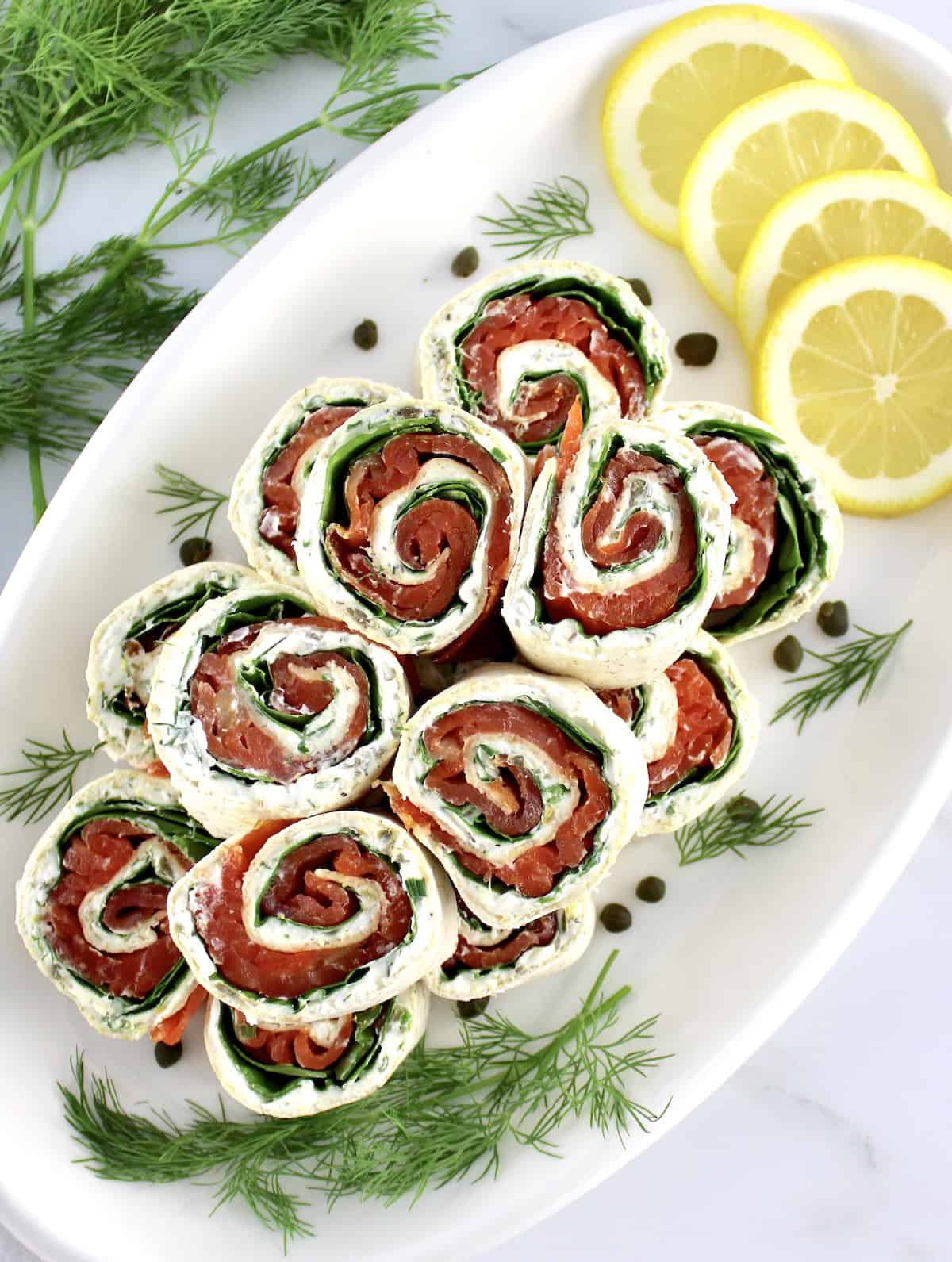 overhead view of Smoked Salmon Pinwheels stacked up on white plate with lemon slices and fresh dill on side