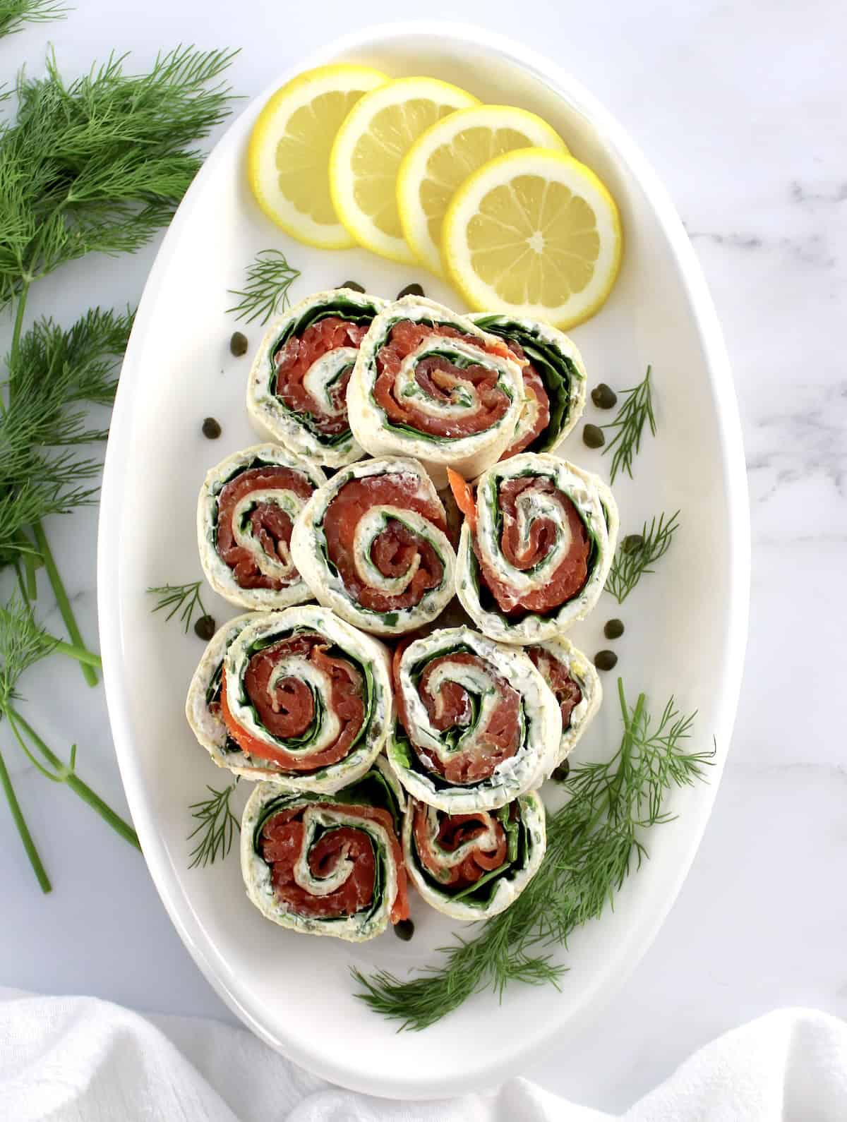 overhead view of Smoked Salmon Pinwheels stacked up on white plate with lemon slices and fresh dill on side