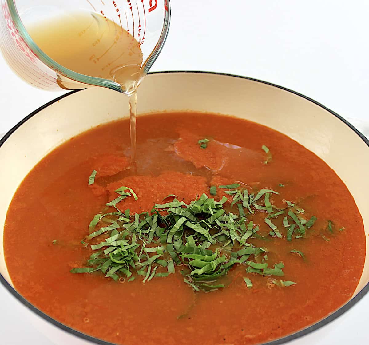 vegetable broth being poured into roasted tomato puree with chopped basil in dutch oven