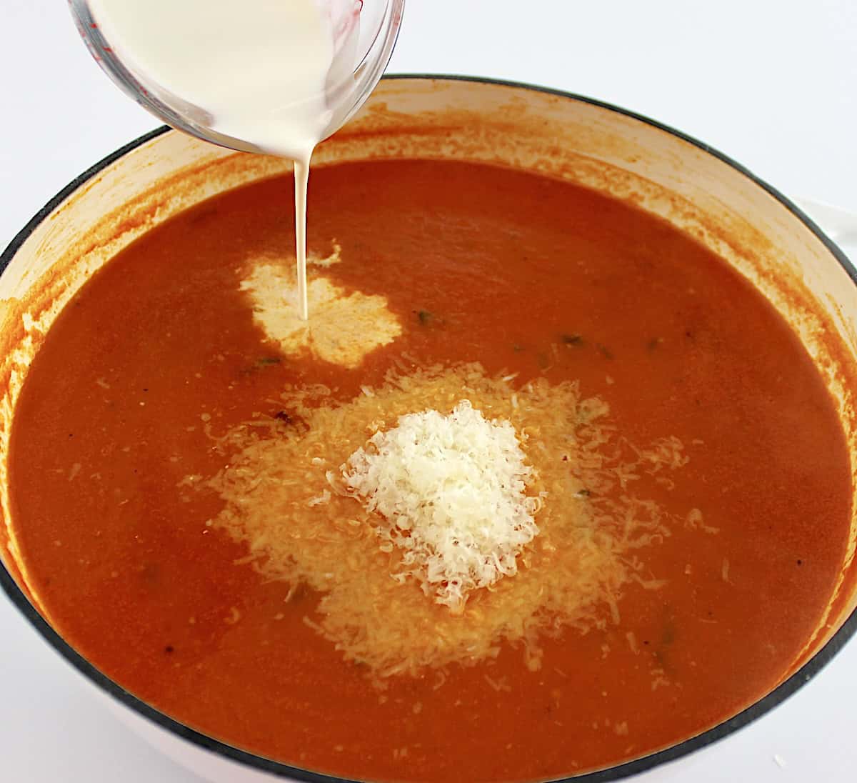 heavy cream being poured into Tomato Bisque with grated parmesan in dutch oven