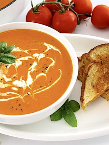 Tomato Bisque in white bowl with grilled cheese, tomatoes and skillet on side