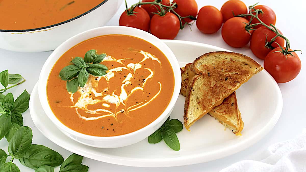 Tomato Bisque in white bowl with grilled cheese, tomatoes and skillet on side