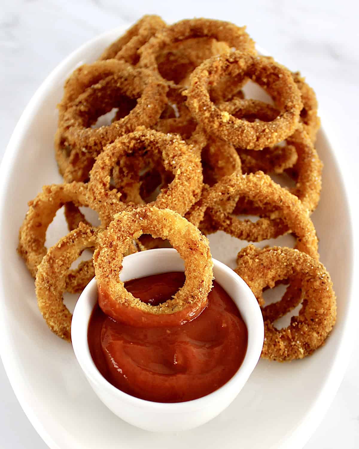 Air Fryer Onion Rings on white plate with one in small white cup with ketchup