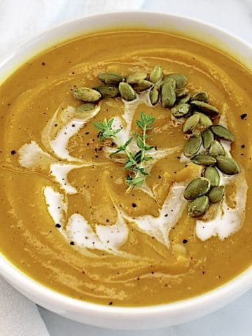 closeup of Butternut Squash Soup in white bowl with cream drizzled on top with pumpkin seeds