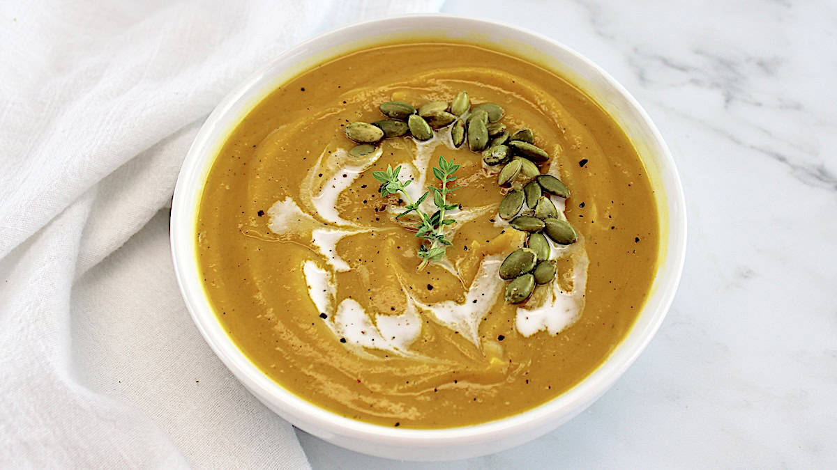 closeup of Butternut Squash Soup in white bowl with cream drizzled on top with pumpkin seeds