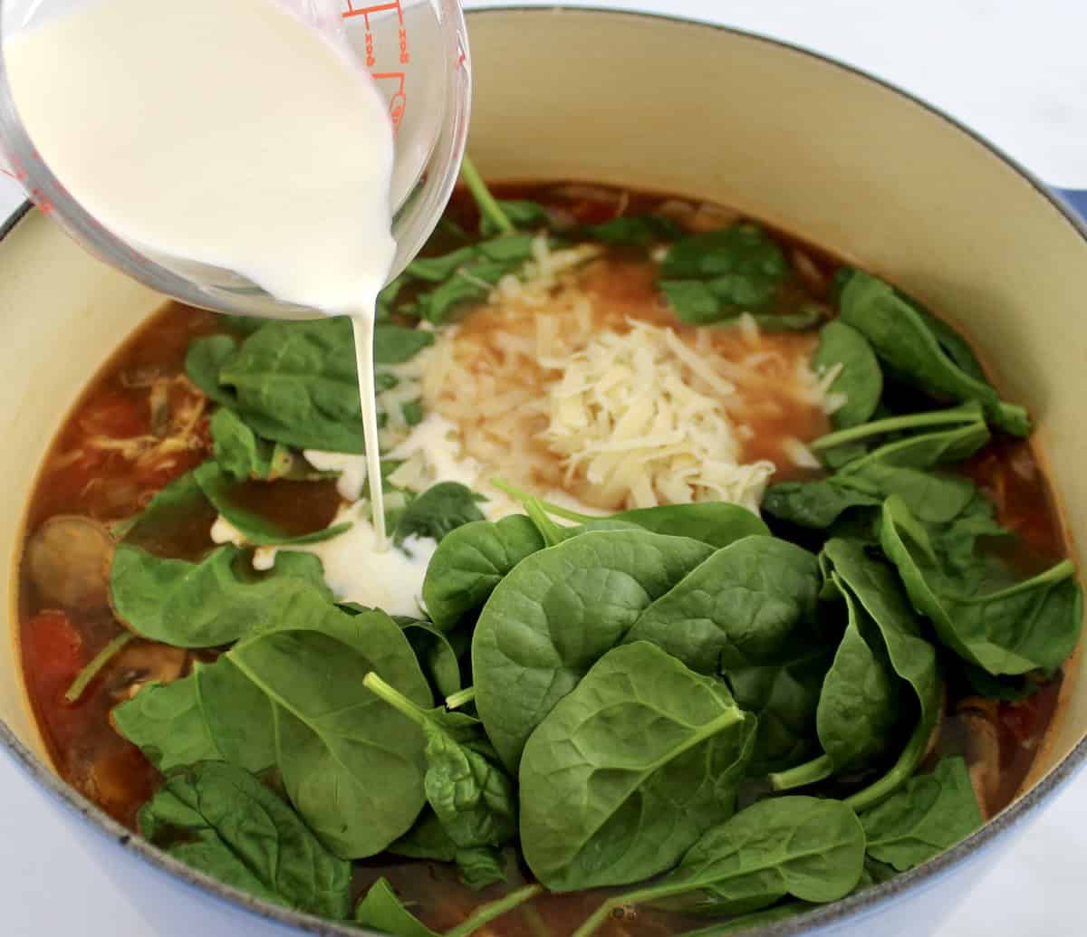 Tuscan Chicken Soup with fresh baby spinach, shredded parmesan and heavy cream being poured in the pot