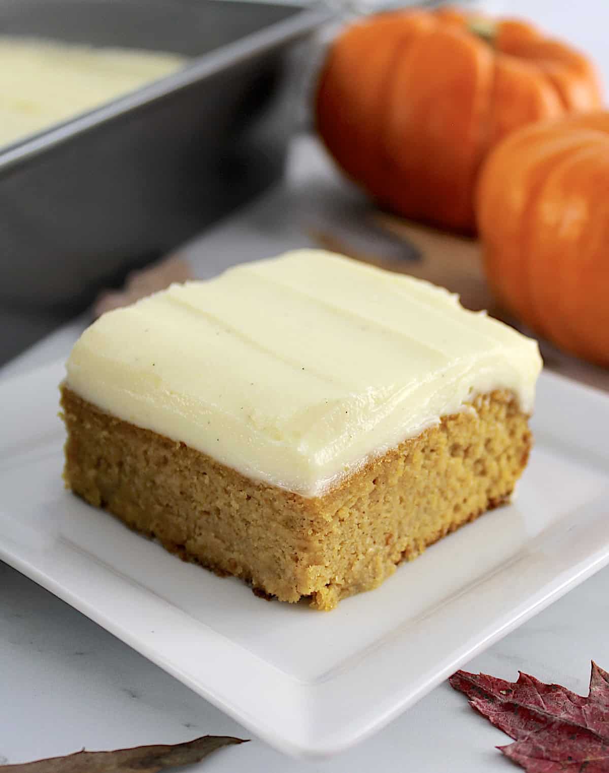 slice of Pumpkin Spice Cake on white plate with pumpkins and baking pan in background