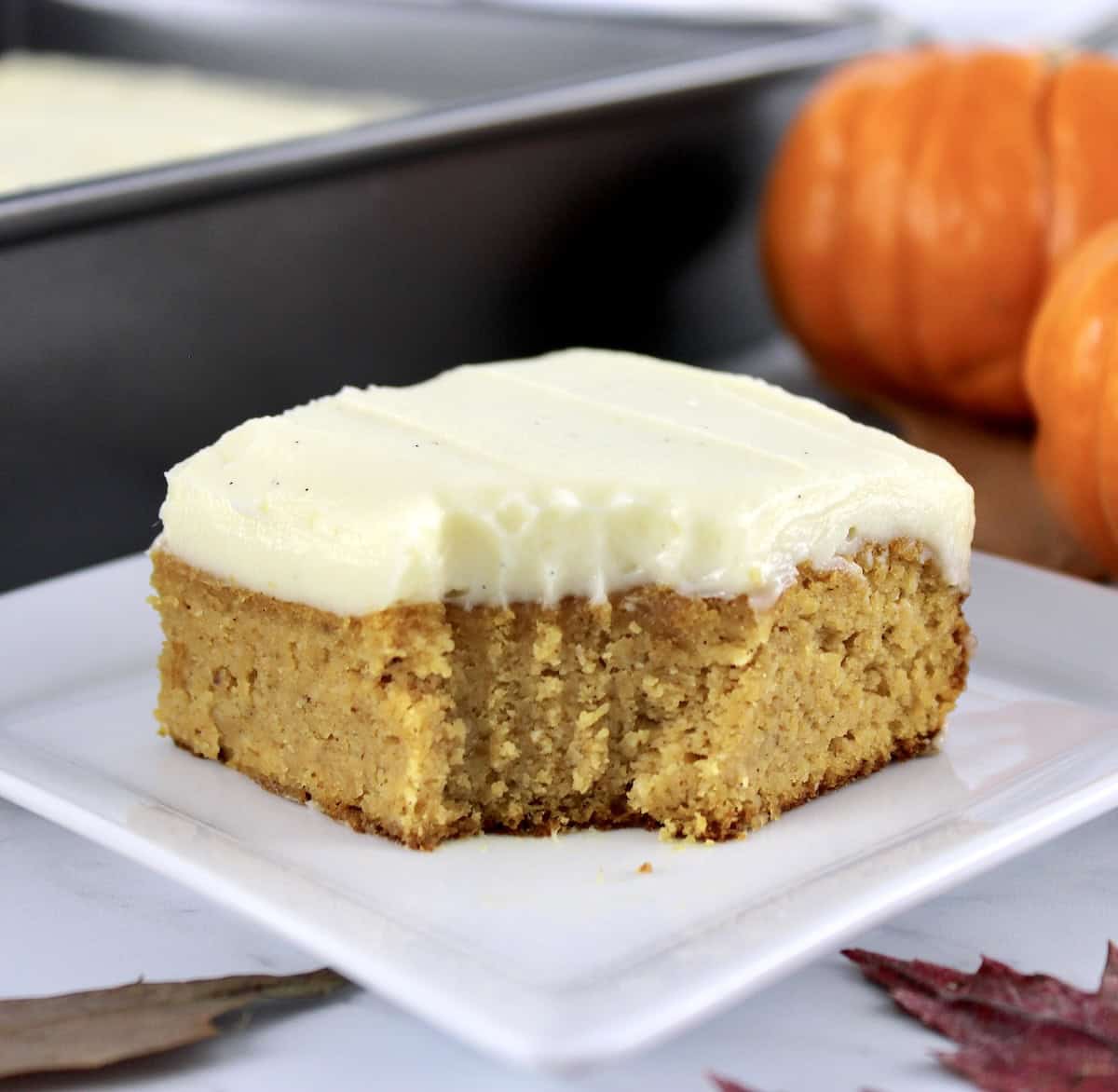 slice of Pumpkin Spice Cake on white plate with bite missing, cake pan and pumpkins in back