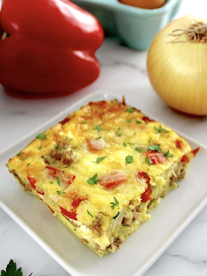 slice of Sausage and Peppers Breakfast Casserole on white plate with red pepper and onion in back