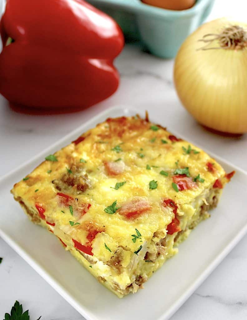 slice of Sausage and Peppers Breakfast Casserole on white plate with red pepper and onion in back