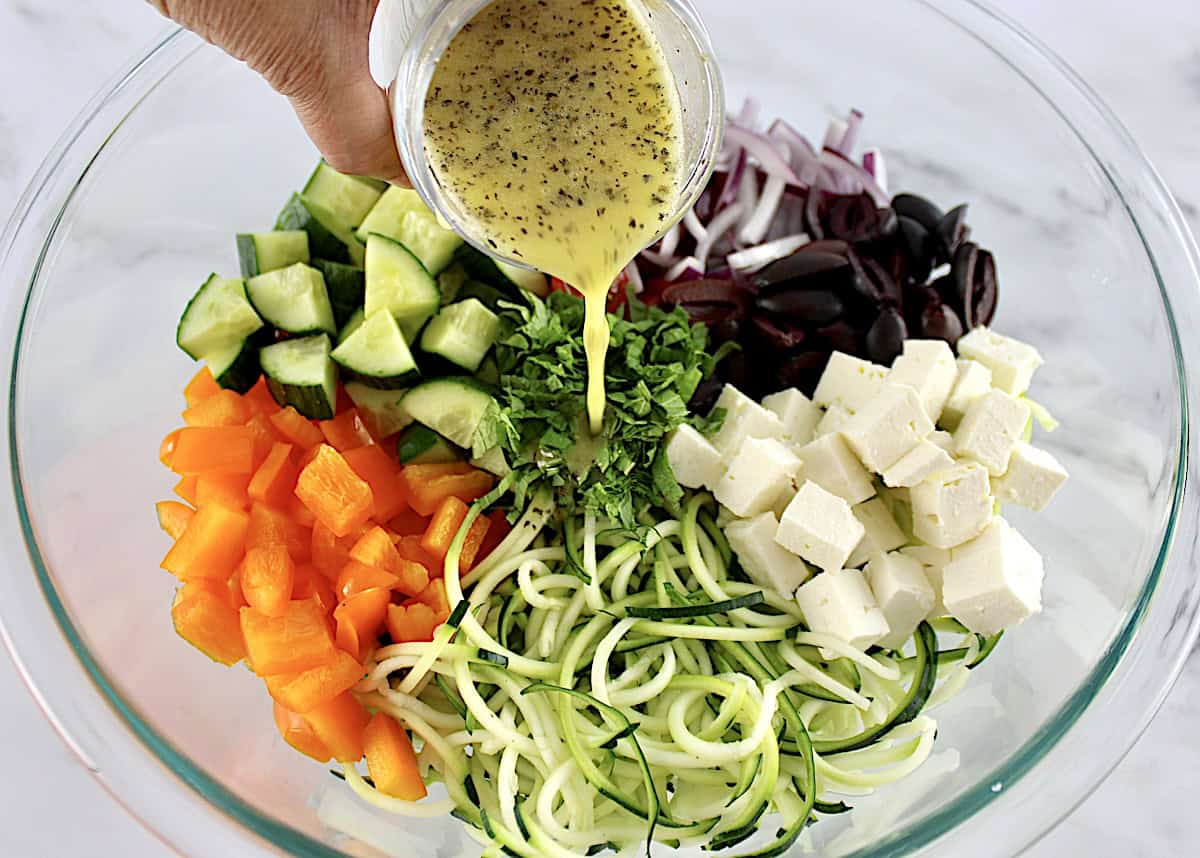 Greek Zucchini Pasta Salad ingredients in glass bowl in sections unmixed with dressing being poured over top