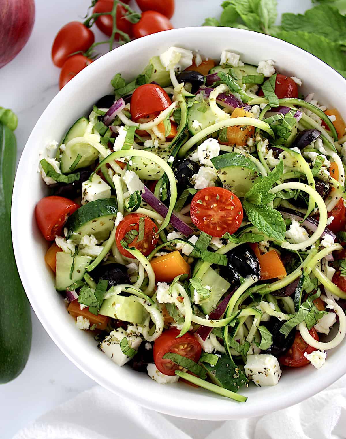 overhead view of Greek Zucchini Pasta Salad in white bowl with veggies in back