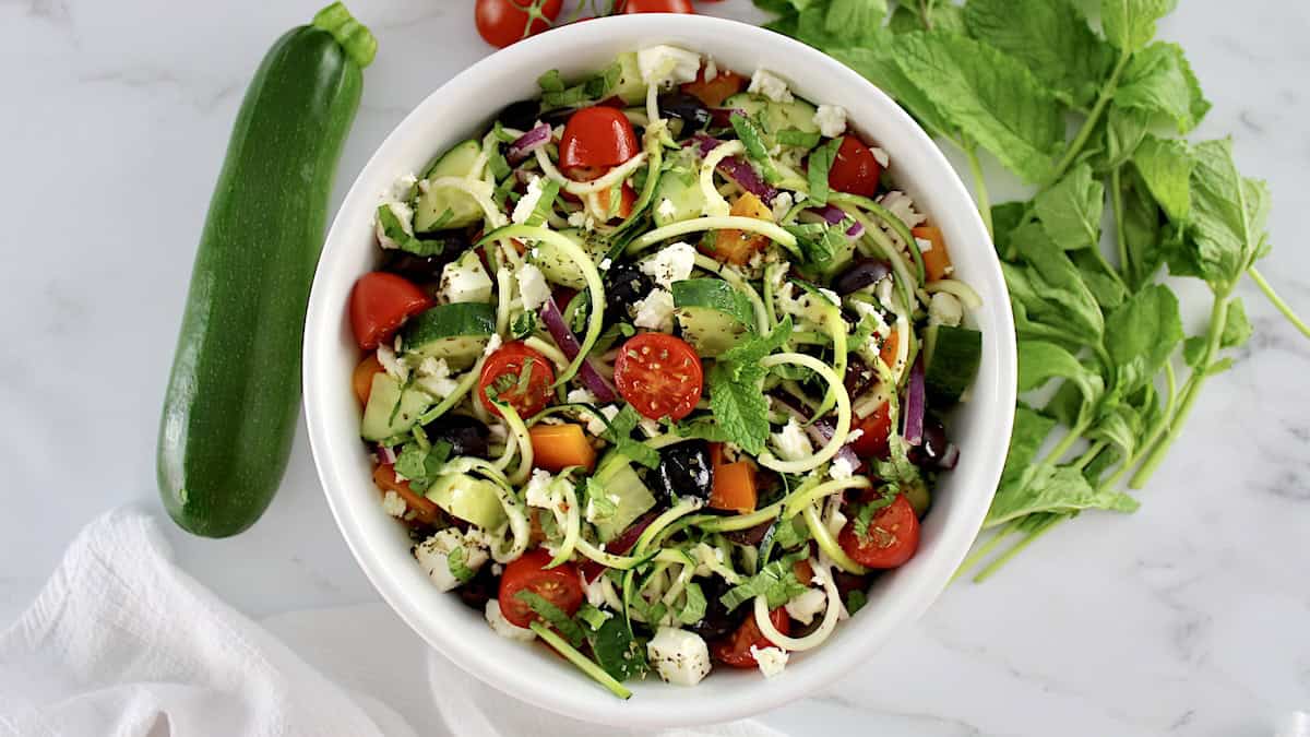 overhead view of Greek Zucchini Pasta Salad in white bowl with veggies in back