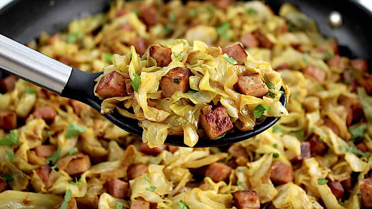 Fried Cabbage with Ham in black serving spoon held up over skillet