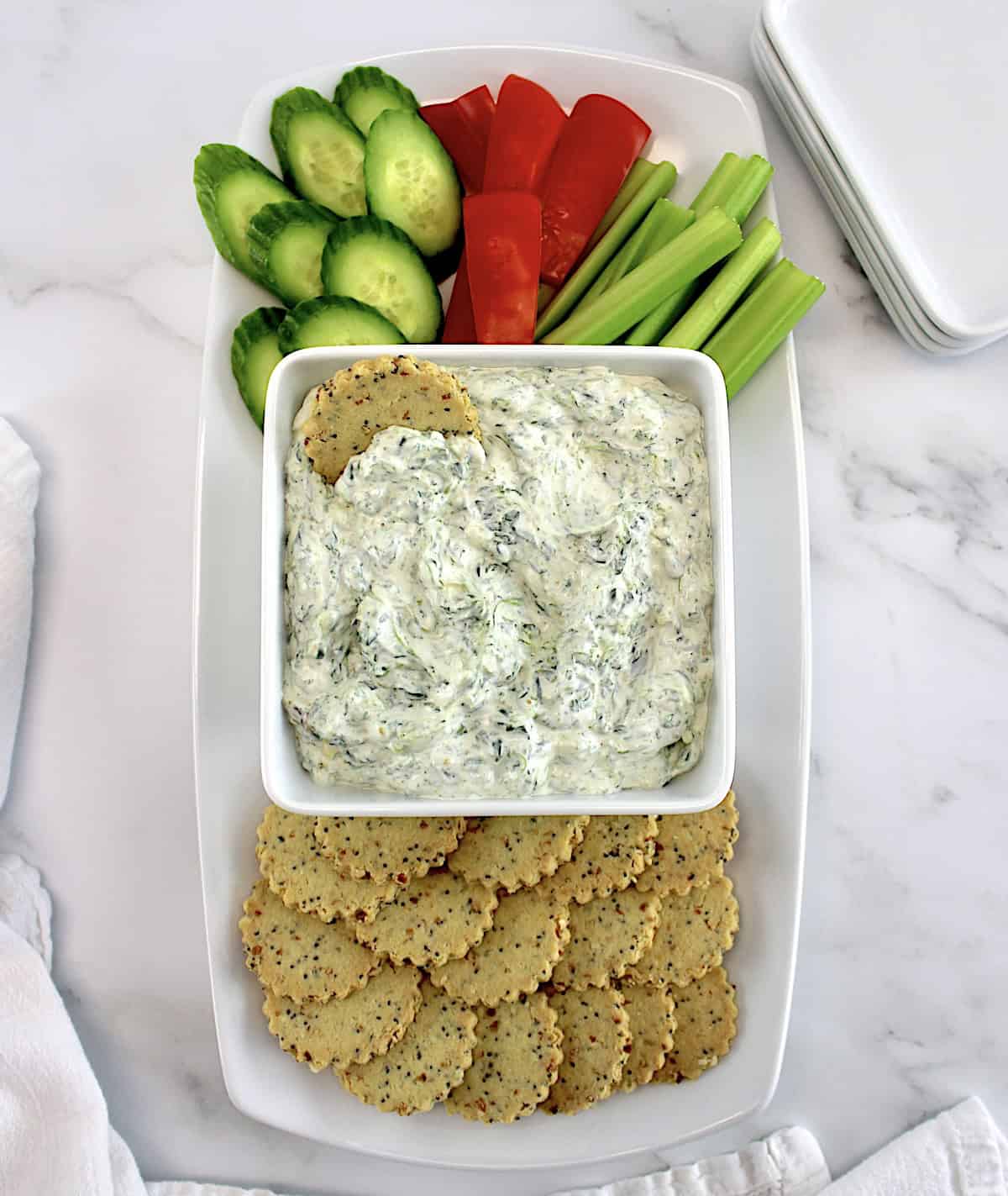 Spinach Dip in white square bowl with crackers, celery sticks, red bell pepper and cucumber slices on side