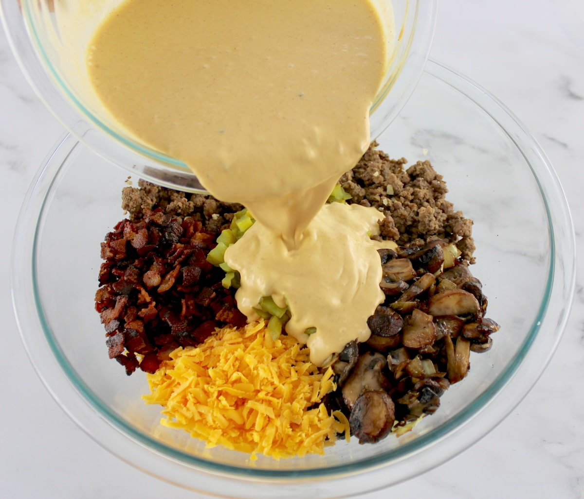 Bacon Cheeseburger Casserole ingredients in glass bowl in sections unmixed with sauce being poured over