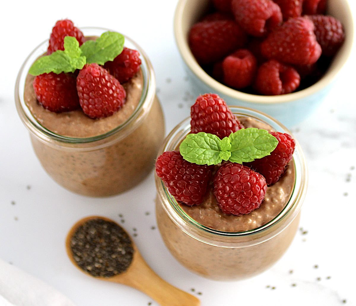 closeup of Chocolate Chia Seed Pudding in 2 open glass jars with raspberries and mint sprig on top