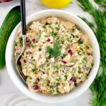 Creamy Dill Salmon Salad in white bowl with fork in it and dill, lemon and cucumbers in back