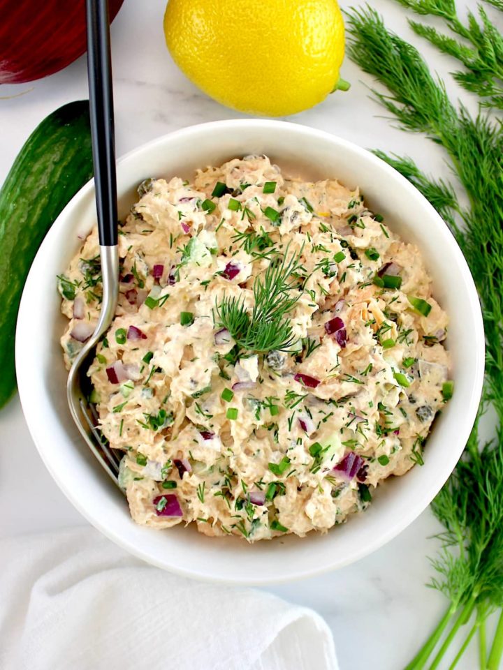 Creamy Dill Salmon Salad in white bowl with fork in it and dill, lemon and cucumbers in back