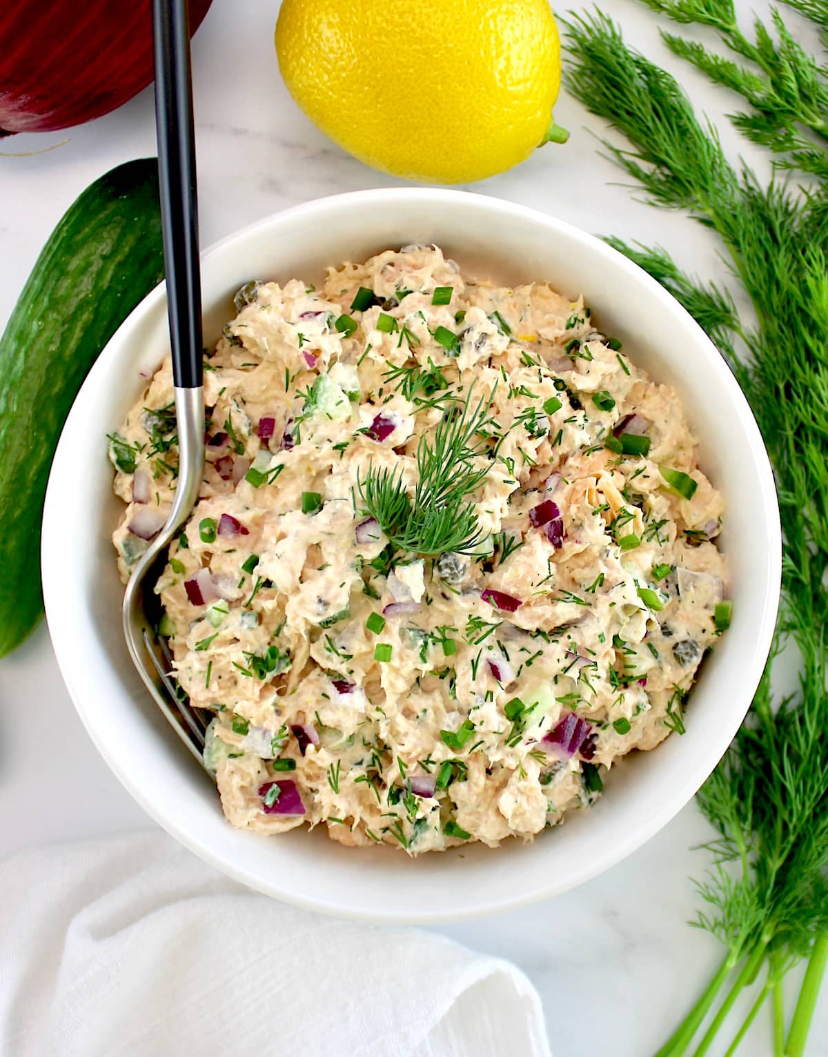 Creamy Dill Salmon Salad in white bowl with fork in it and dill, lemon and cucumbers in back