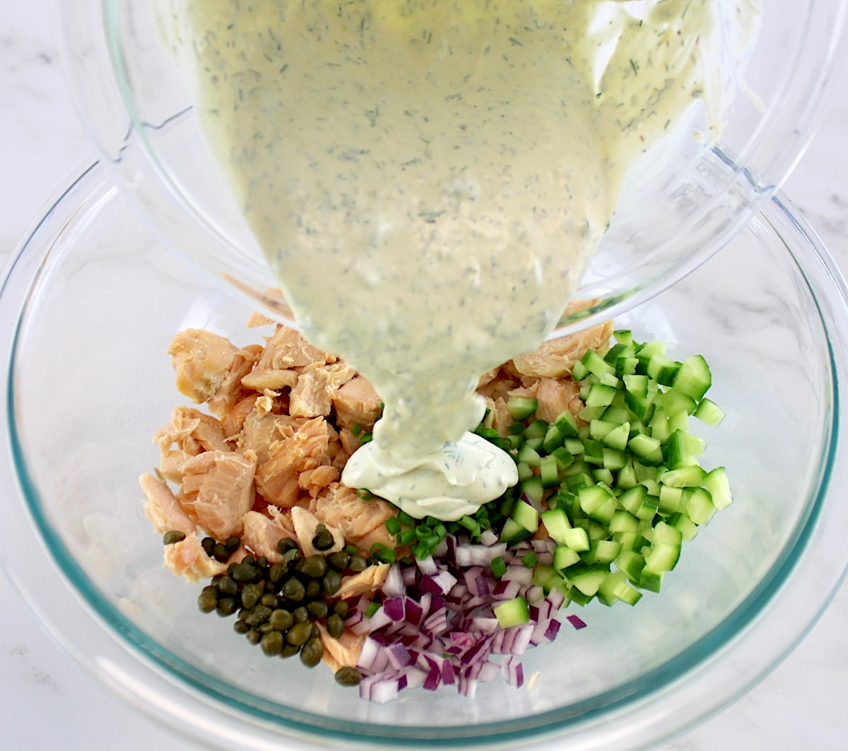 flaked salmon, diced cucumber, red onion, capers and chives in glass bowl unmixed with creamy dill sauce pouring over top