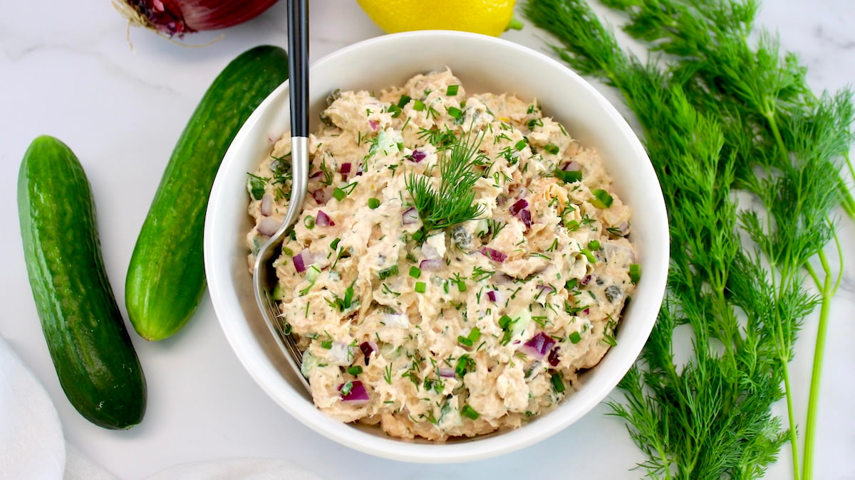 overhead view of Creamy Dill Salmon Salad in white bowl with fork in it and dill, lemon and cucumbers in back