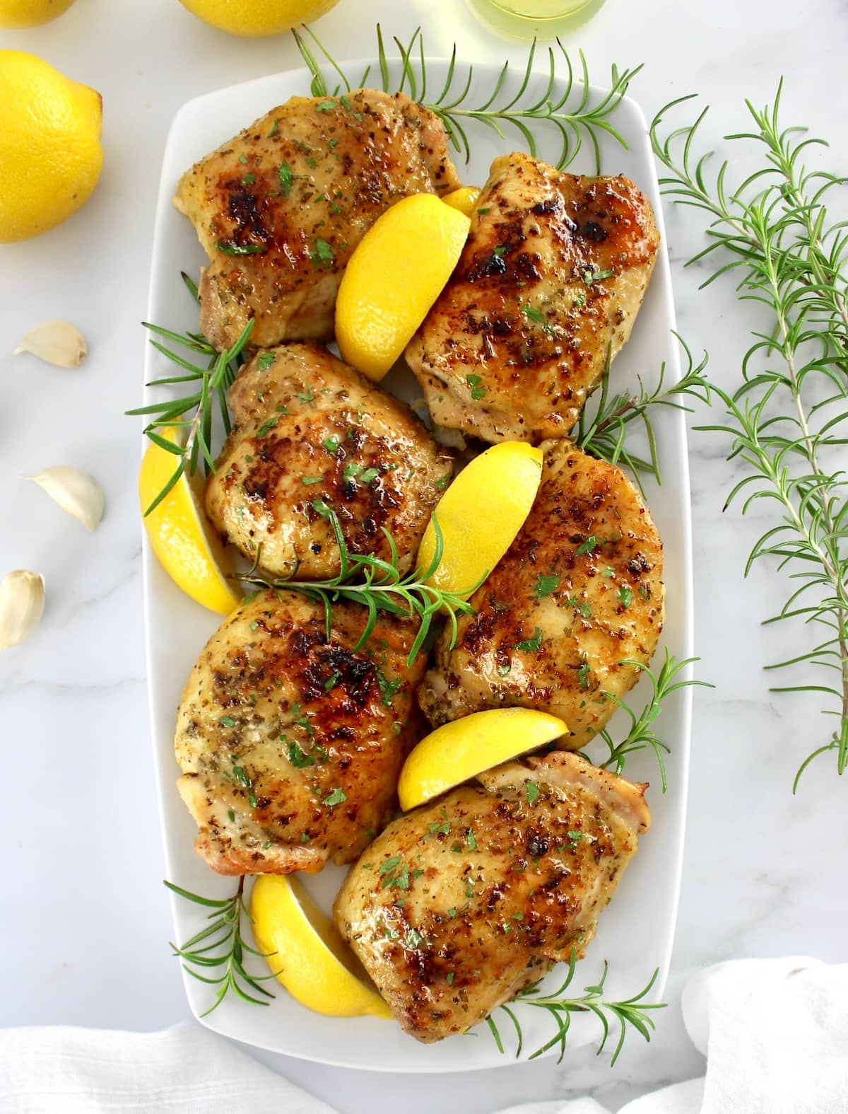 6 pieces of Greek Lemon Chicken thighs on white plate with lemon wedges and rosemary