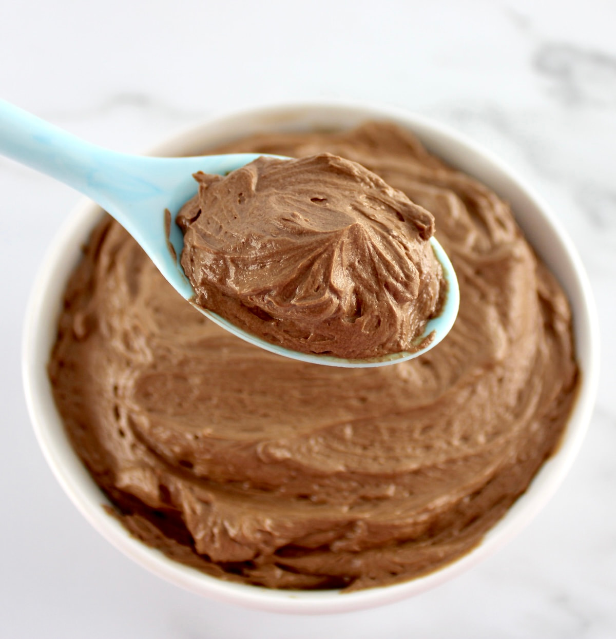 closeup of Keto Chocolate Frosting in blue spoon held up over white bowl of most frosting