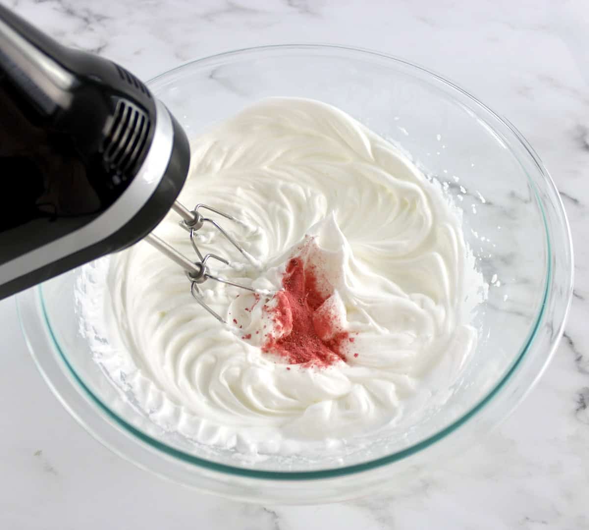 egg whites with strawberry powder in glass bowl being whipped with hand mixer