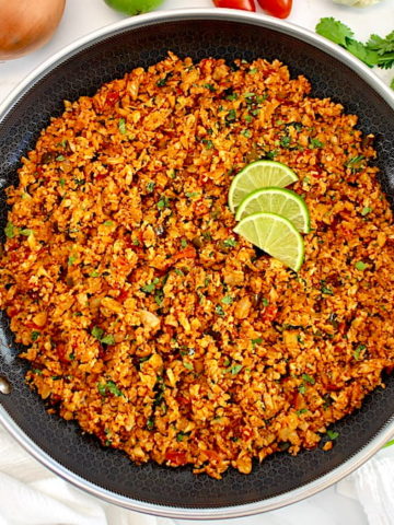 overhead view of Mexican Cauliflower Rice in skillet with lime slices and veggies in back