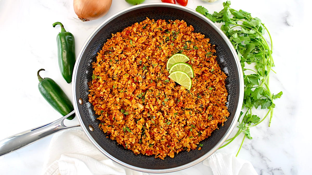 overhead view of Mexican Cauliflower Rice in skillet with lime slices and veggies in back