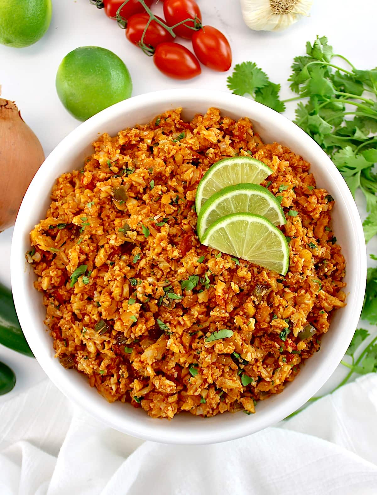 overhead view of Mexican Cauliflower Rice in white bowl with 3 lime slices and veggies in back