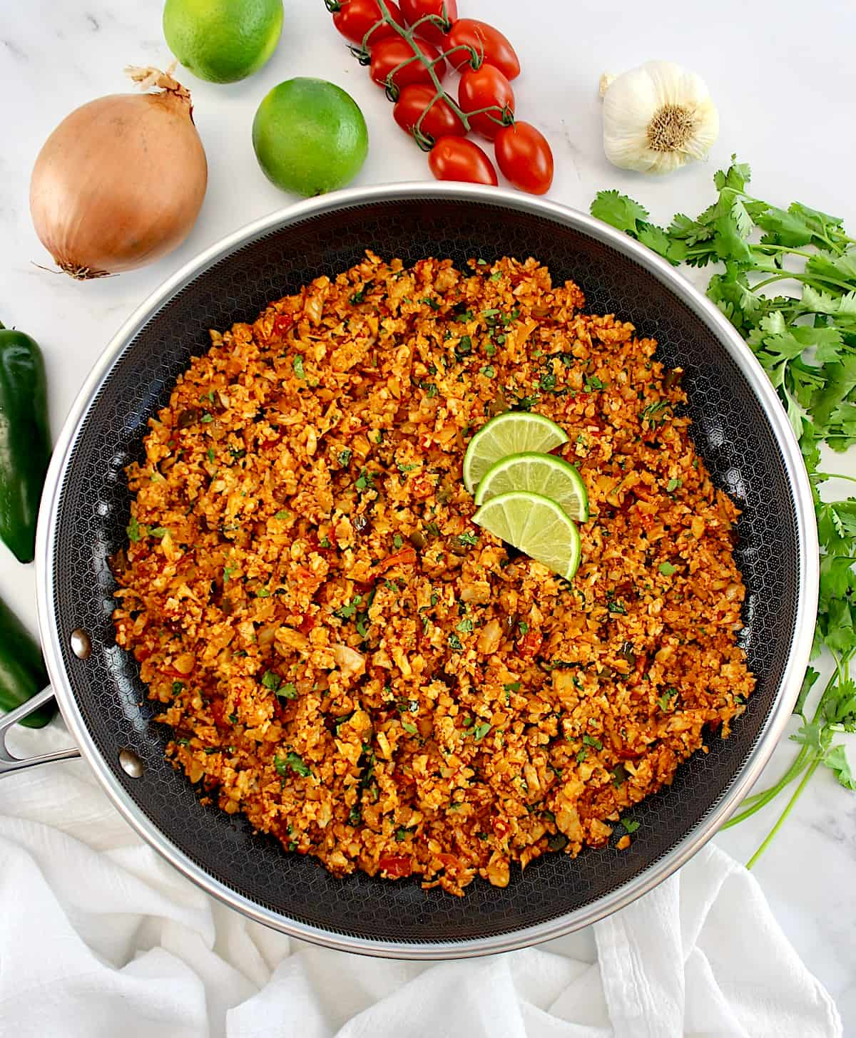 overhead view of Mexican Cauliflower Rice in skillet with lime slices and veggies in back