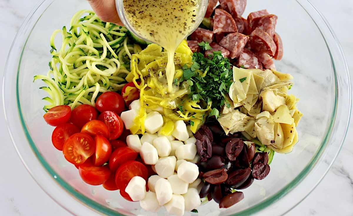 Zucchini Noodle Pasta Salad ingredients in glass bowl in sections unmixed with dressing being poured over top